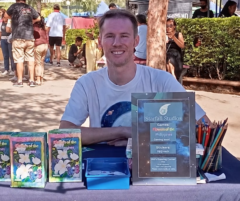 Jamie at a market table with card games, colored pencils, and a starry studio banner.