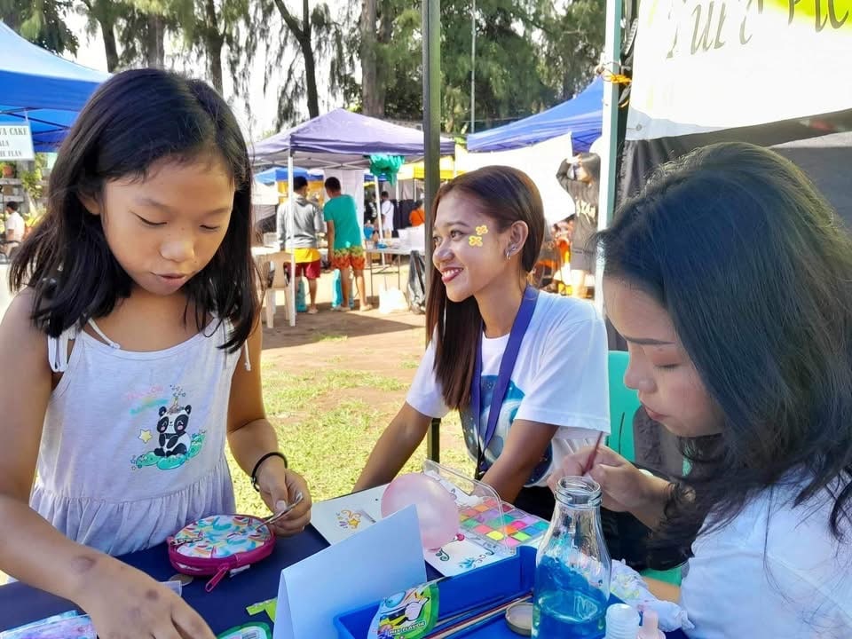 A girl draws at a market table while two volunteers help.