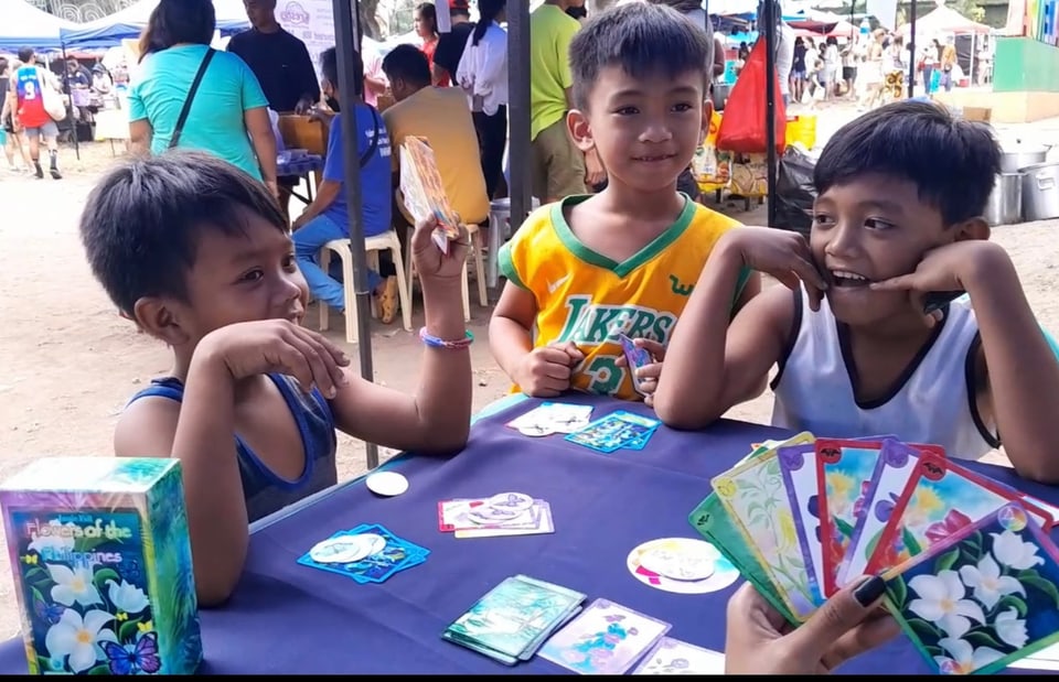 Three boys at a table playing a card game, one fanning out flower cards.