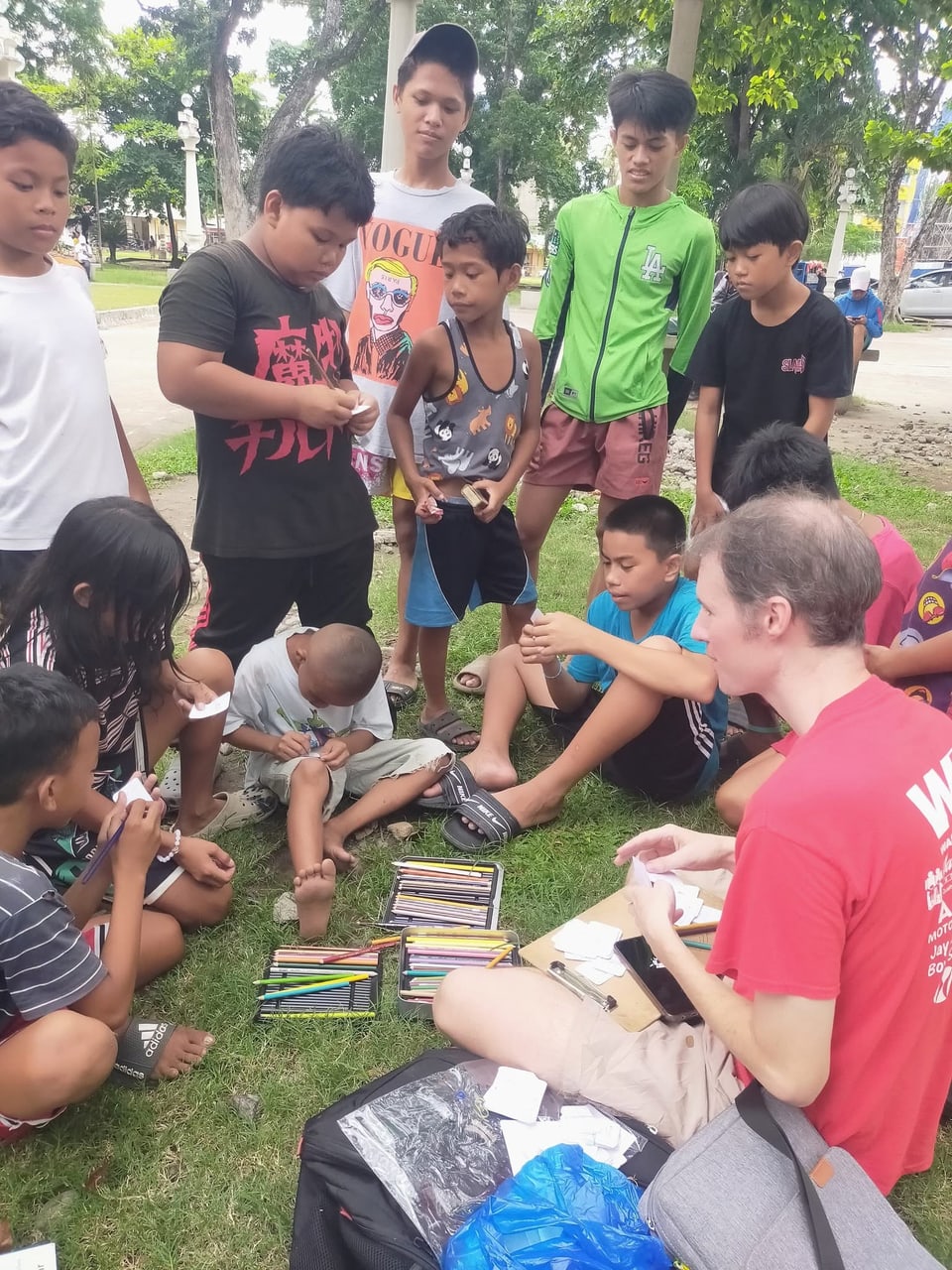 Children crowd around a table of colored pencils at an outdoor drawing session.