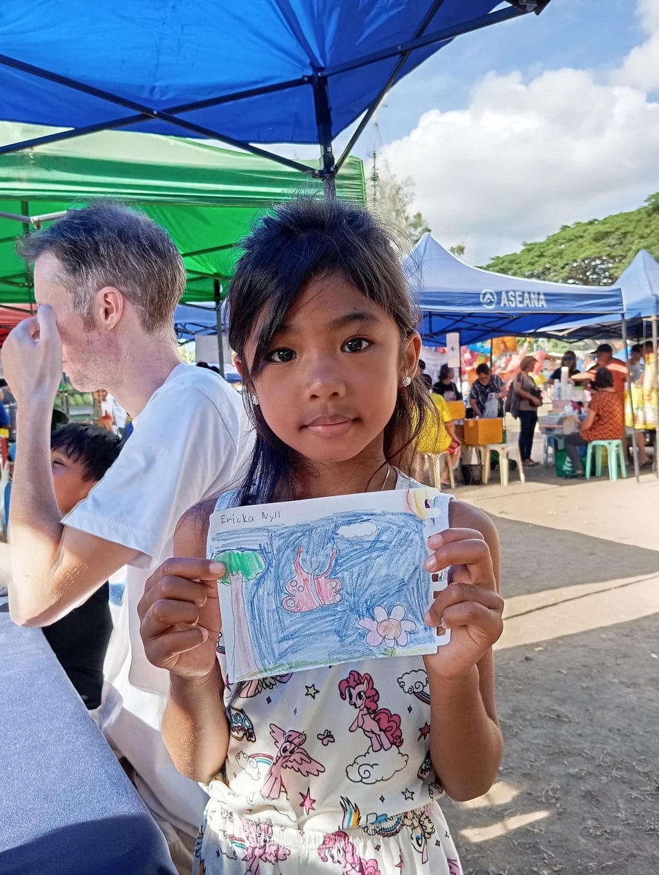 A girl holds up her drawing at an outdoor market.