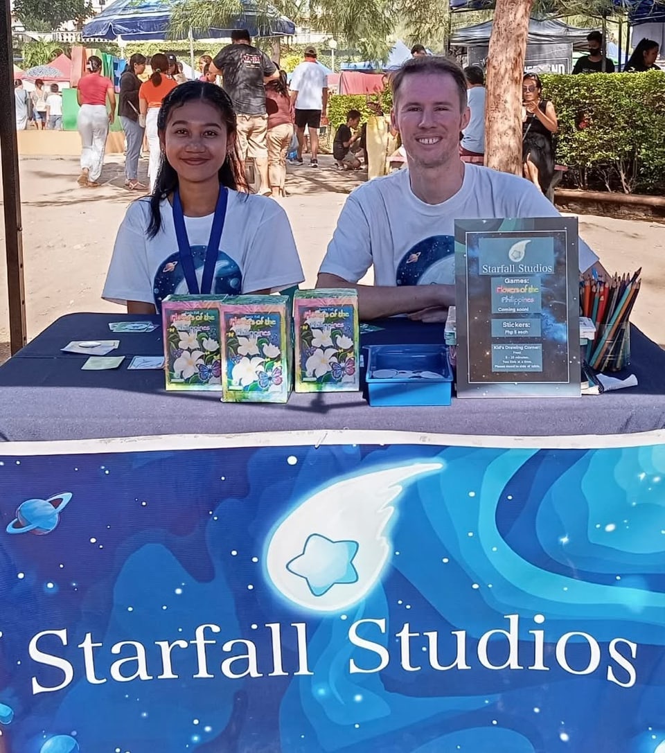 Two people at a market table with card games, stickers, and a starry studio banner.
