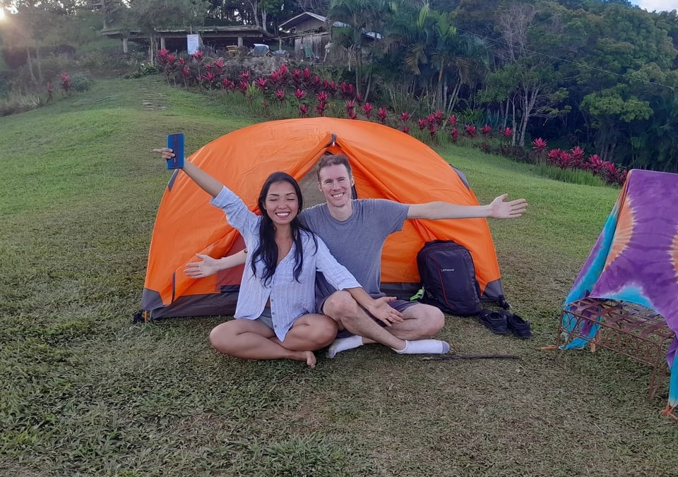 Two people sit in front of an orange tent on a green hillside.