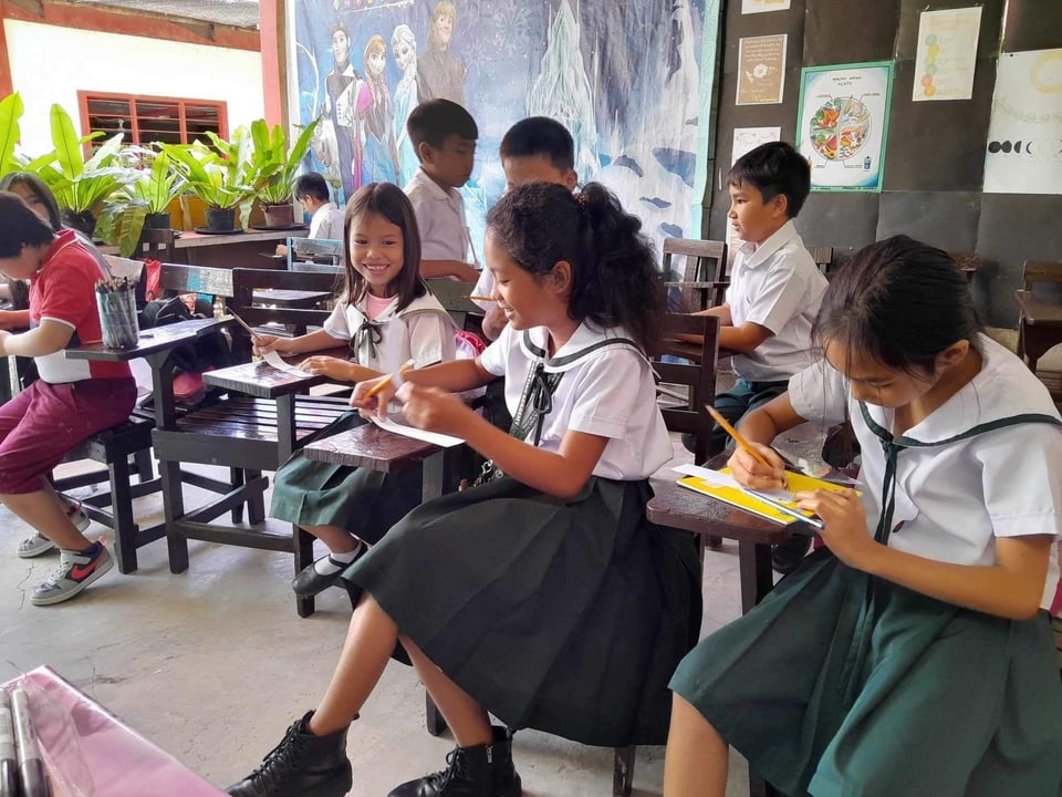 Students draw together at wooden desks in a classroom.