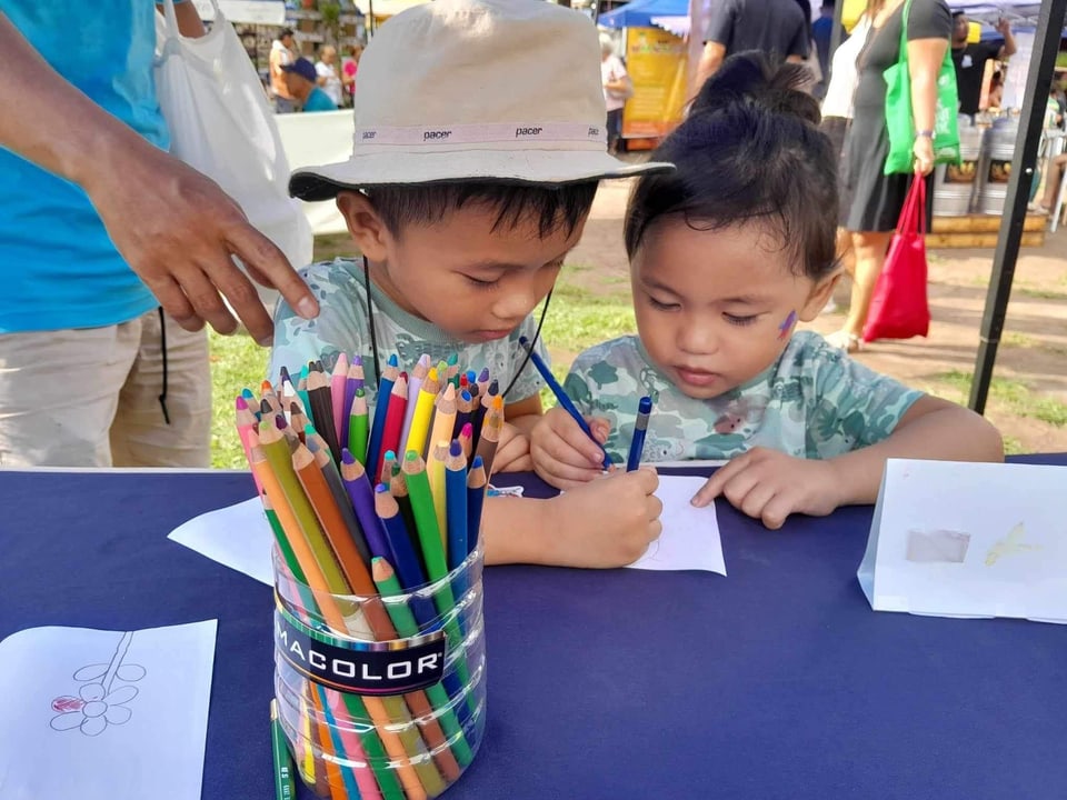 Two small children color at a table with a cup of pencils.